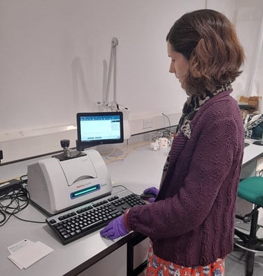 Photograph showing a short-haired woman in a purple jumper standing while working at a desk. She is reading results from a small machine while typing on a keyboard.