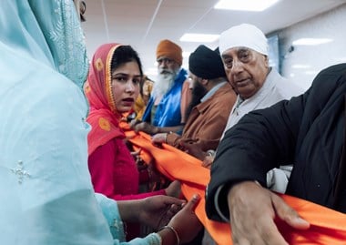 People line either side of a length of orange fabric, looking along it as they fold it.