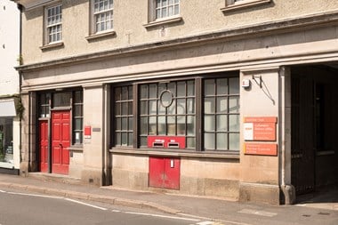 The facade of a two story stone building with wooden multi-paned windows and a red, panelled front door. Above there are three Georgian-style sash windows. There are post boxes set into the front of the building.