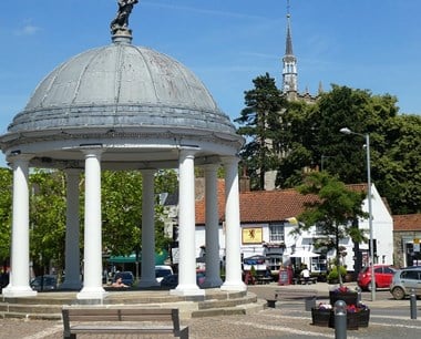 Domed structure resting on seven white pillars. In the background is a two-storey white-painted pub with sign that reads: Red Lion.