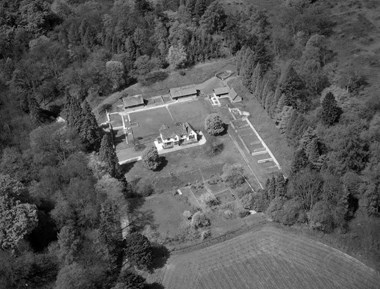 Black and white oblique aerial photograph of an archaeological site. In the centre is a house-like building, sent in lawns surrounded by lines of excavated walls marking the positions of former buildings. The ground is terraced and sloped to the front of the house, leading to grassland. To the sides and rear of the site are areas of woodland. 
