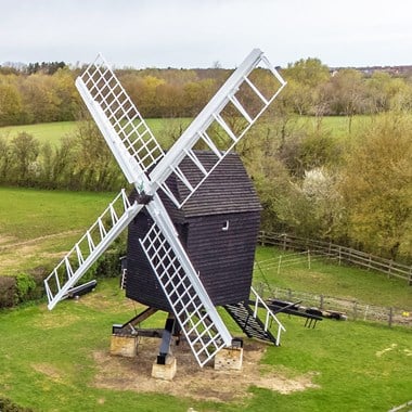 High-angle photograph of a black wooden trestle windmill with white vanes in a green field. 