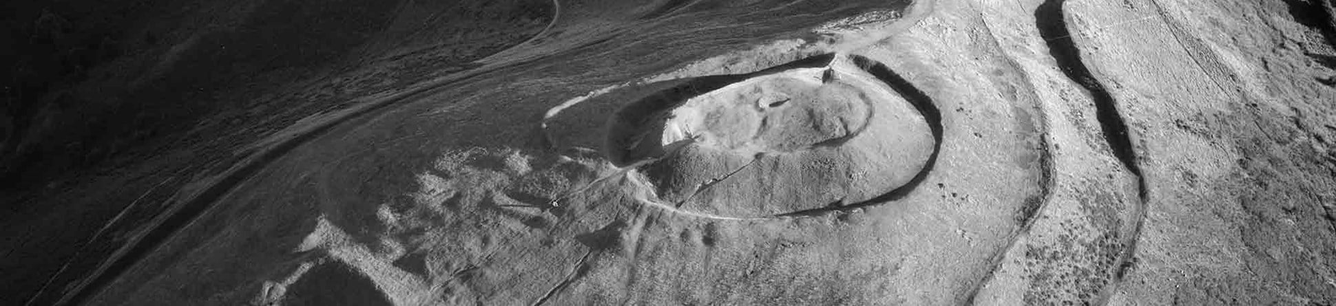 Black and white oblique aerial photograph of a univallate Iron Age hillfort, photographed in low sunlight that casts strong shadows, revealing banks, ditches and tracks surrounding a central, circular mound. The hillside drops steeply to the foreground and either side of the narrow spine of the hill.