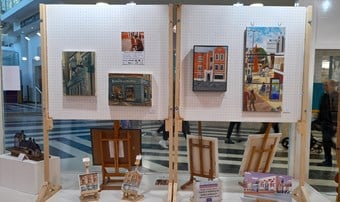 Artworks mounted on standing display boards. Beneath them, other artworks stand on small easels, facing out through the shop window into the shopping centre, where pedestrians' legs can be seen passing by.