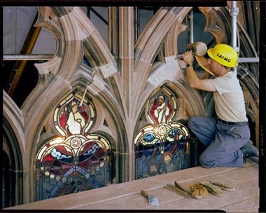 A stonemason in yellow Laing hardhat works on stone around stained glass at the Carlisle Cathedral.