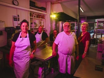 Five people stand in pink light around a kitchen steel work surface wearing aprons. 