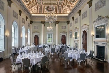 An interior photograph of tables and chairs set up for a wedding in an elegant Georgian-era function room, with a chandelier, decorative columns and a decorative ceiling
