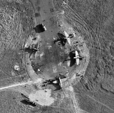 A black and white aerial photograph showing four aircraft parked at an aifield hardstand. The hardstand is surrounded by ground that is marked with lines of vehicle tracks.