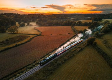 A photograph of a steam train passing through the countryside at sunset.