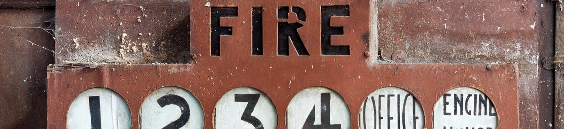 An old, red alarm panel attached to a wall. Underneath the word FIRE are indicators reading 1, 2, 3, 4, Office, Engine House.