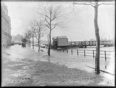 Black and white photo of River Thames flooding the Embankment and buildings along it. A horse and cart are being driven towards the viewer in the middle distance.