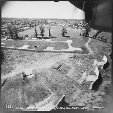 A black and white oblique aerial photograph taken at low level. In the foreground are rifle ranges with canted walls. In the middle ground is a track with parking areas. In the background, beyond a line of spaced trees, is a recreation field between the buildings of a military camp.