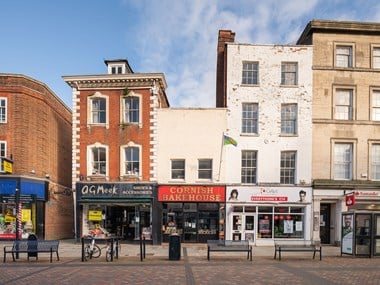 A view across a high street to a row of three old buildings. Each is a different height and made of different materials. The red-brick building on the left has fine stonework details and a sign reading 'Meeks Shoes.' It is closed down and looks in poor repair.