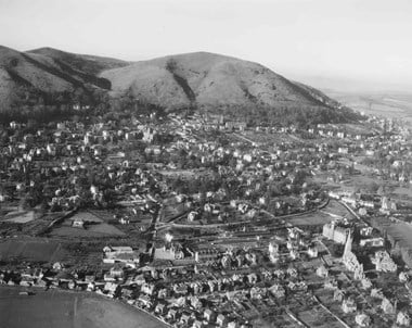Black and white oblique aerial photograph showing a multitude of buildings of a town situated at the base of a range of undulating hills. Much of the town is well spread out, with trees and open spaces lining roads and separating well-spaced detached and semi-detached houses. The houses stand out, illuminated by low sunlight. Many of them have large front and rear gardens that frame long shadows. A railway line runs across the foreground and several church towers and steeples can be seen amongst the trees and houses.