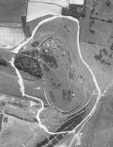 A black and white vertical aerial photograph of a rounded rectangular enclosure bounded by fields and open countryside. Surrounding the site is a light-coloured, irregular line marking some kind of boundary feature.