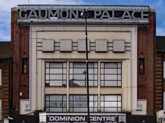 A three-storey high street art deco cinema. The "Gaumont Palace" "Dominion Centre" with pedestrians passing outside. 