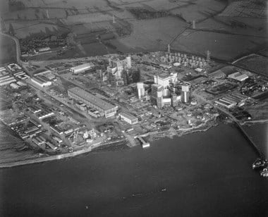 Black and white oblique aerial photograph of a construction site adjacent to a stretch of river. Much of the bottom section of the photograph is calm water. The central section features the construction site of a nuclear power station, including the erection of two reactor buildings. Beyond the construction site are fields bordered with hedges and trees. Parallel lines in the fields suggest evidence of ridge and furrow ploughing. 