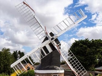 A wooden windmill with sail frames atop a brick base. Additional buildings in background and to the sides.