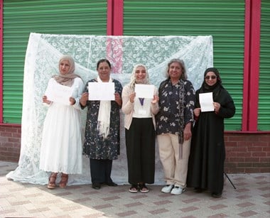 Five women stand in a line, four of them holding a piece of paper each with handwritten words: "Yeah, I did it!" They stand in front of closed shutters and a temporary backdrop of white net curtain fabric. 