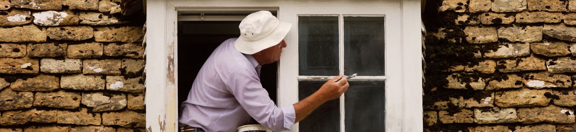 A man leaning out of a dormer window of an old cottage to paint it. 