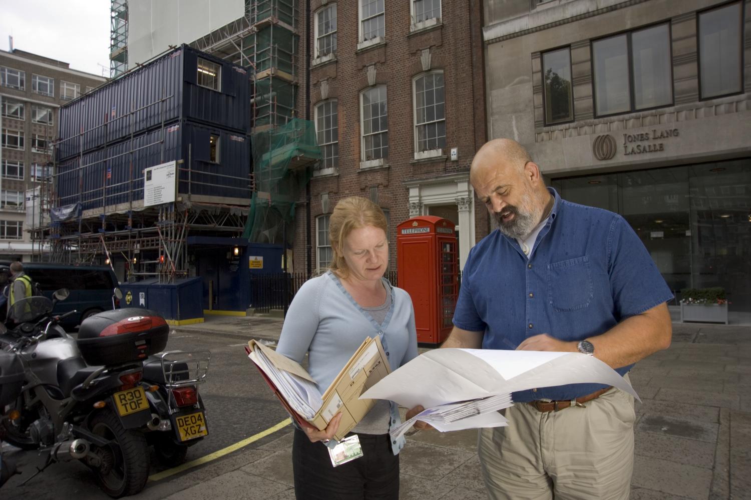 Two people reviewing documents on a city street outside an office building. Construction scaffolding and a red telephone box are visible in the background.