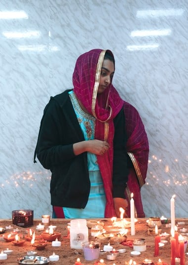 A woman in traditional costume stands in front of a sandpit filled with different burning candles.