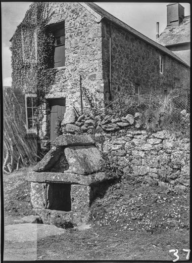 Black and white photograph showing a squat, stone structure with sides, top and pitched roof made from slabs of granite. It is on a grass verge in front of a stone wall. In the background is a two-storey stone farm building. The left edge of the photograph shows evidence of double-exposure.