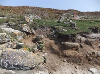 A photograph of an archaeological site on a beach with low walls, part of the structure has been washed away