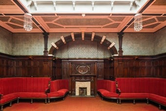 Red velvet benches line wood-panelled walls. Above the panelling, the walls are covered in ceramic tiling. In the centre of the image is an arched recess with a marble fireplace and wood-panelled mantelpiece.