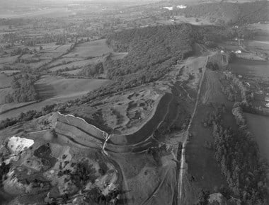 Black and white oblique aerial photograph of a prehistoric earthwork on a hilltop. Curving banks and ditches dominate the centre of the image, enclosing a triangular area featuring hollows and mounds. Fields and woodland extend into the distance.