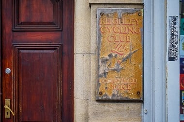 A peeling sign with embossed lettering reading "Keighley Cycling Club Ltd - Registered Office" next to a dark wooden door. 