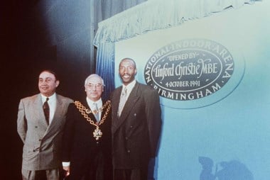 Three men pose for a picture and all of them wear suits. In the middle, the man wears an ornamental necklace showing he is a mayor. In the background there is circular sign on a blue background a sign saying ‘Linford Christie MBE. Birmingham’. In the bottom left of the image, there is the shadow of a photographer.