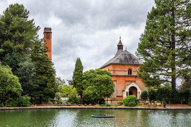 A large red-brick building and tower stand beyond a body of water, surrounded by conifer trees. 