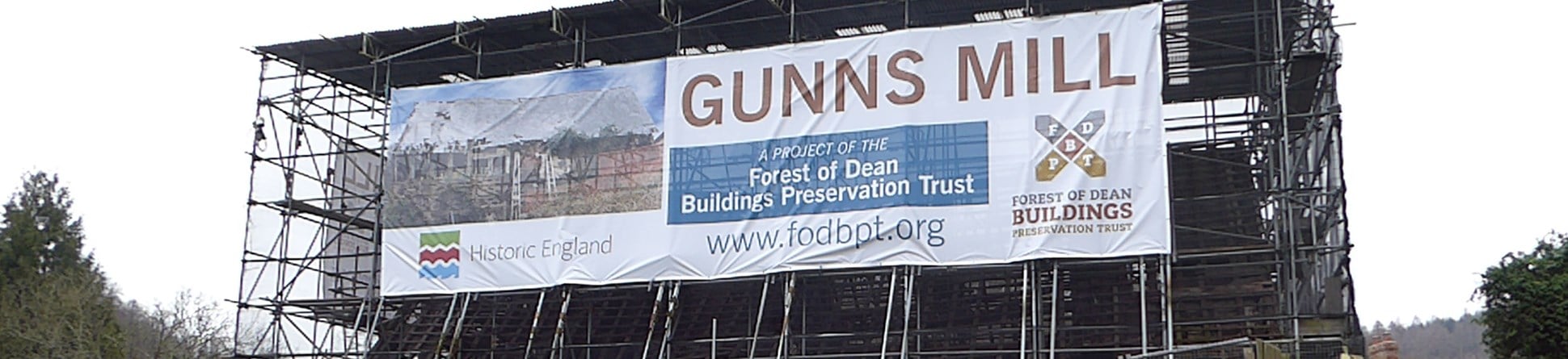 A photograph of a historic mill building entirely protected with scaffolding, showing a banner with the words 'Gunns Mill'.
