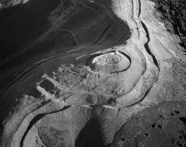 Black and white oblique aerial photograph of a univallate Iron Age hillfort, photographed in low sunlight that casts strong shadows, revealing banks, ditches and tracks surrounding a central, circular mound. The hillside drops steeply to the foreground and either side of the narrow spine of the hill.