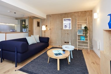 A photo of the sitting area of a modern flat, with a blue sofa, coffee table, and a bare concrete wall in the background.