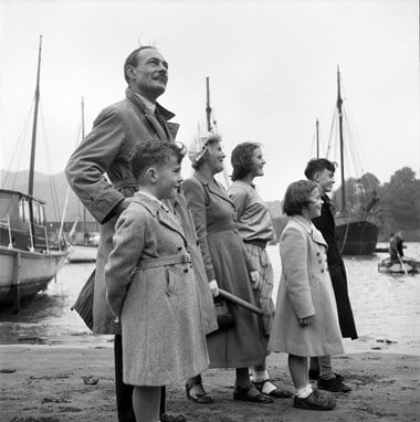 A side view of a man, women and children posed on a beach, with boats in the water beyond