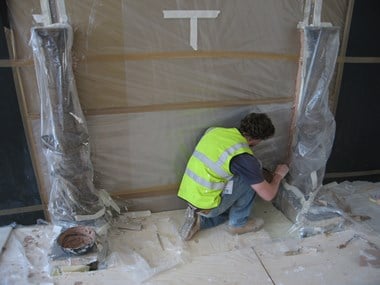 A man in a high-viz vest kneels in front of a wall covered in boards and plastic sheeting.