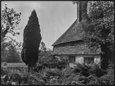 Black and white photograph showing an oblique view of an elevation of a farmhouse. The building is partly obscured by trees and bushes. The lower level of the farmhouse is faced in stone. Above it is tile-hung. A tall, brick chimney protrudes through a steeply-pitched roof. To the left of the farmhouse is a timber gate and beyond is a field and trees.