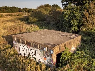  A large square concrete building is seen from above, surrounded by scrub and greenery. Vivid graffiti can be seen on the front of the building. 