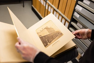 A photo album being handled by a member of staff in the Historic England Archive store. The gloved hands of the member of staff carefully hold open pages of the album. The facing page shows a photographic print of buildings on one side of a street.