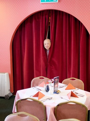 A man's head poking out between long velvet curtains, with restaurant chairs and tables in the foreground.