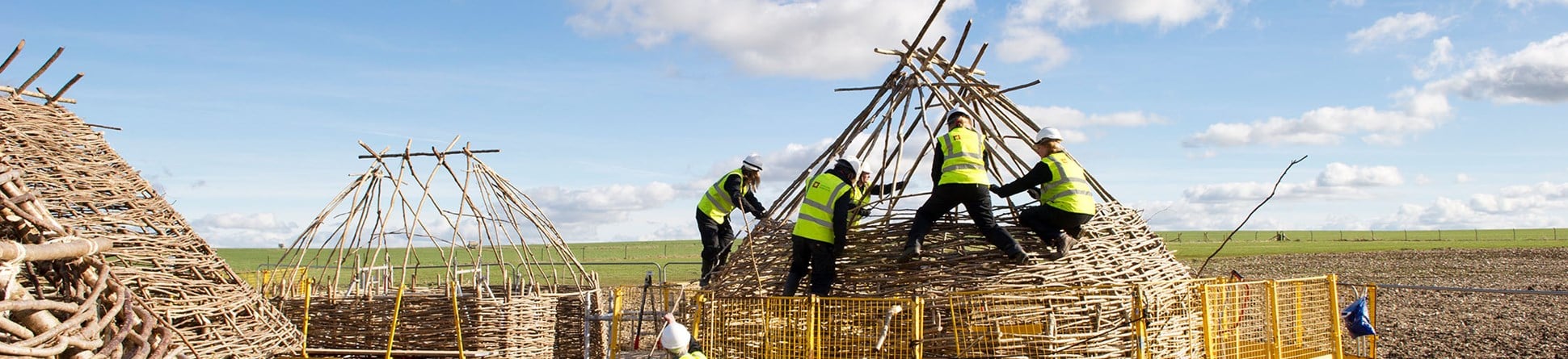 Five people in high-visibility clothing and helmets constructing a Neolithic-style hut.