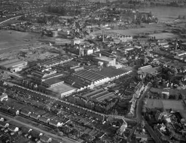 Black and white oblique aerial photograph of a large factory complex surrounded on three sides by streets with terraced and semi-detached housing and industrial buildings. The factory features one- and two-storey buildings with parallel rows of saw-tooth roofs, and flat-roofed blocks of two and three storeys. Tall chimneys extend from several industrial sites in the photograph. To the rear of the factory is a patch of muddy or sandy ground adjacent to a stretch of river. Beyond the river are fields and a further expanse of town.