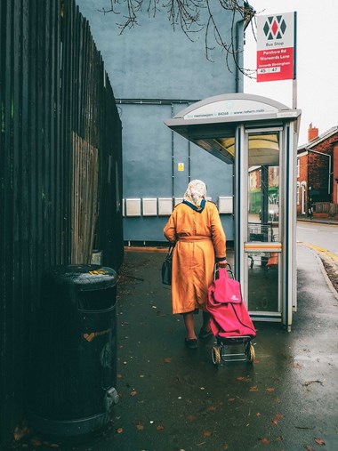 A woman wearing a headscarf, pulling a shopping trolley behind her, heading for the bus stop.