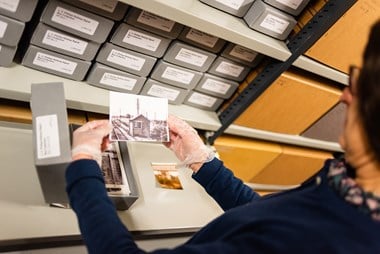 A colour photograph being handled in the Historic England Archive store. The gloved hands of the member of staff carefully holds a photo of a small brick building next to a railway line. In the background are shelves with cardboard boxes. 