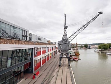 A photograph showing a large, grey metal crane on a dockside. Beside it is a long industrial building named M Shed. 