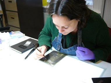 Photograph showing a dark-haired woman in gloves and an apron working at a desk. She is using a paintbrush to work on a piece of glass on the tabletop.