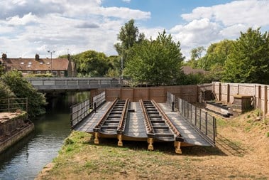 A clean low metal structure alongside a canal channel, with a modern rail bridge in the background. 