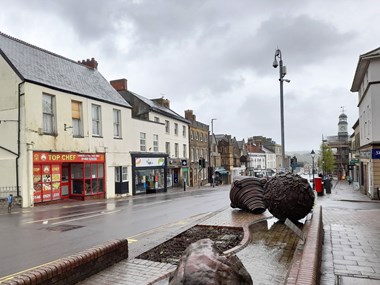 A historic high street on a wet day. In the foreground is a block of possibly sandstone and two large artworks of rusted metal.